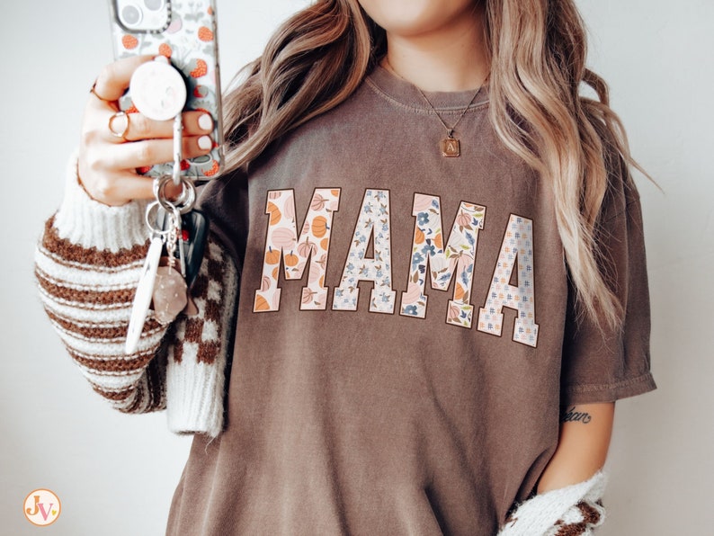 May include: A brown t-shirt with the word "MAMA" printed in large, white letters. The letters are decorated with a floral pattern in shades of pink, blue, and orange.