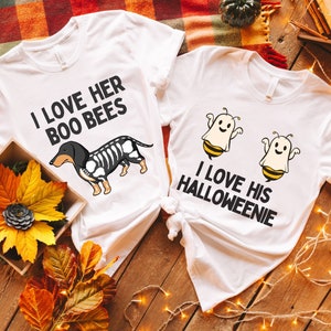 May include: Two white t-shirts with a Halloween theme. One shirt says "I love her boo bees" and has a black and white dachshund skeleton with bee wings. The other shirt says "I love his Halloweenie" and has two ghosts with bee wings.