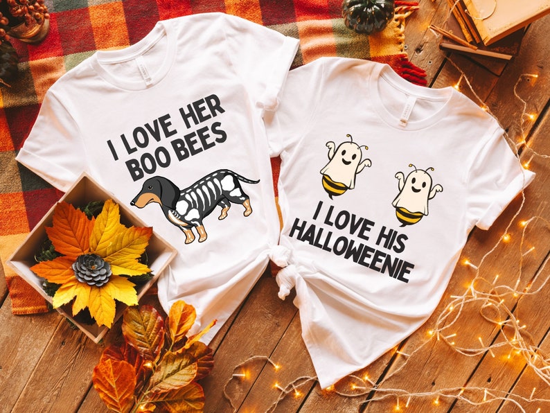 May include: Two white t-shirts with a Halloween theme. One shirt says "I love her boo bees" and has a black and white dachshund skeleton with bee wings. The other shirt says "I love his Halloweenie" and has two ghosts with bee wings.