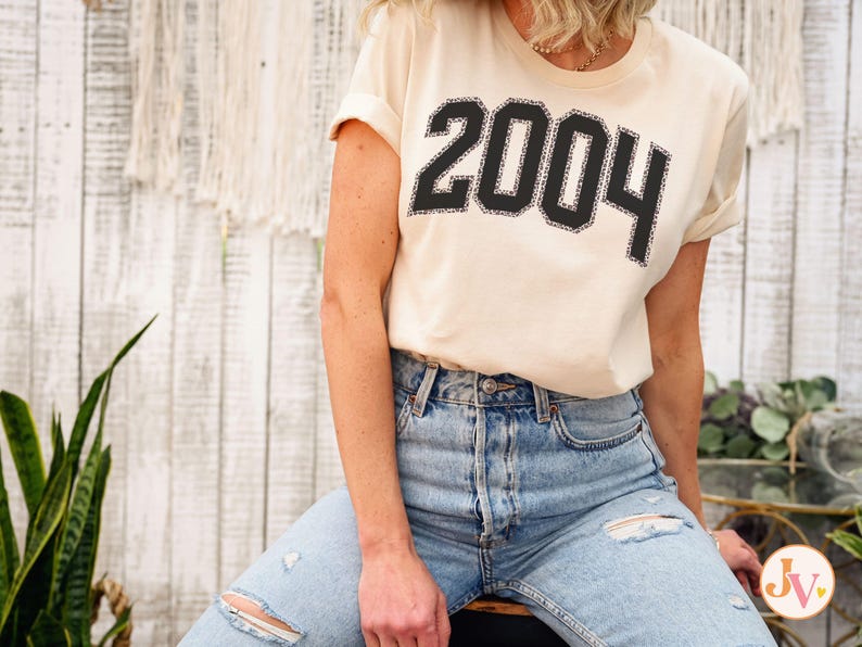 May include: A woman wearing a cream colored t-shirt with the year 2004 printed in black. She is wearing light blue jeans with rips in the knees.