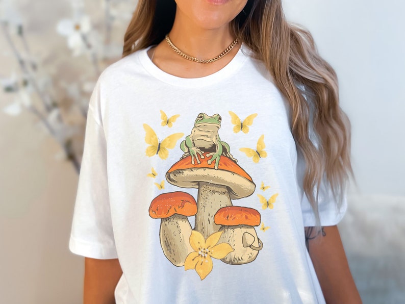 May include: White t-shirt with a green frog sitting on a mushroom with yellow butterflies around it.