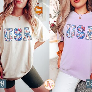 May include: Two women wearing t-shirts with the word "USA" spelled out in floral letters. The shirt on the left is ivory and the shirt on the right is orchid.