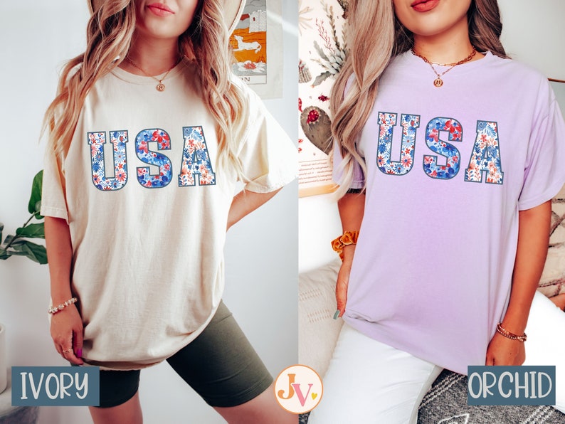 May include: Two women wearing t-shirts with the word "USA" spelled out in floral letters. The shirt on the left is ivory and the shirt on the right is orchid.