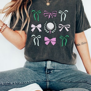 May include: Dark gray t-shirt featuring a golf ball on a tee surrounded by a wreath, and several bows in shades of green, pink, and white. The shirt is paired with blue jeans and white sneakers. The overall theme is golf.