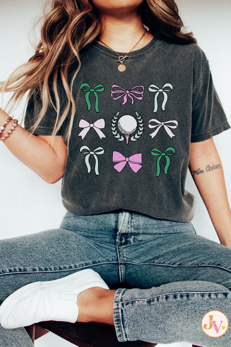 May include: Dark gray t-shirt featuring a golf ball on a tee surrounded by a wreath, and several bows in shades of green, pink, and white. The shirt is paired with blue jeans and white sneakers. The overall theme is golf.