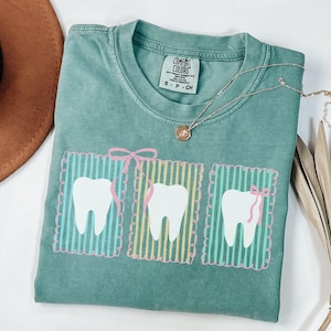 May include: A light green Comfort Colors t-shirt featuring three white teeth designs with pink bows and striped backgrounds. A silver necklace with a small pendant is draped on the shirt. A brown hat is visible in the upper left corner.