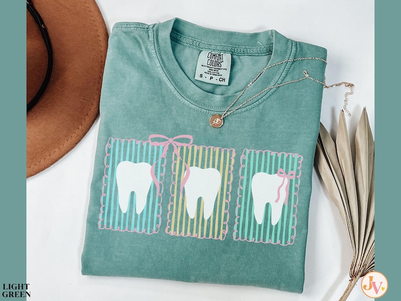 May include: A light green Comfort Colors t-shirt featuring three white teeth designs with pink bows and striped backgrounds. A silver necklace with a small pendant is draped on the shirt. A brown hat is visible in the upper left corner.