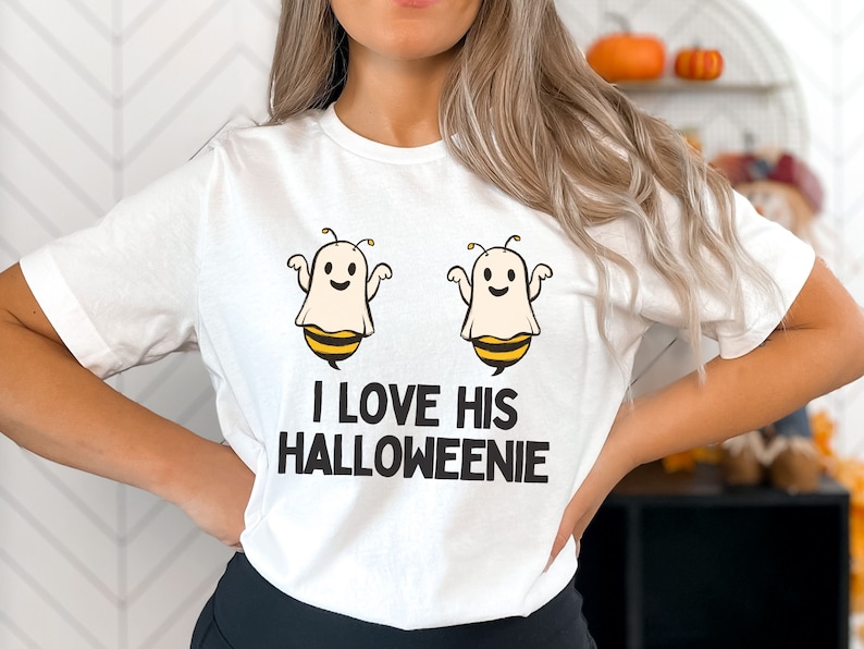 May include: White t-shirt with a graphic of two ghosts dressed as bees. The text "I love his Halloweenie" is printed below the ghosts.