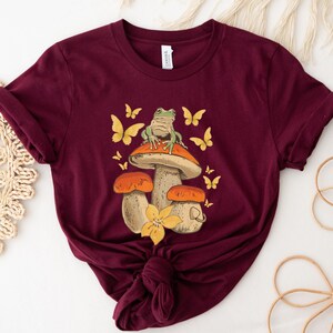 May include: A maroon t-shirt with a graphic of a green frog sitting on a mushroom with orange and yellow accents. The mushroom has a white stem and is surrounded by butterflies.