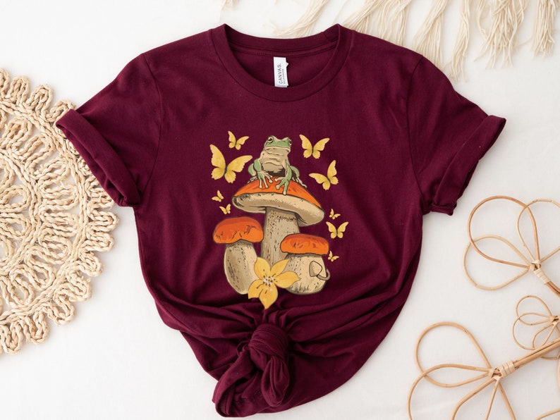 May include: A maroon t-shirt with a graphic of a green frog sitting on a mushroom with orange and yellow accents. The mushroom has a white stem and is surrounded by butterflies.