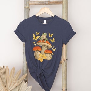 May include: A navy blue t-shirt with a graphic of a green frog sitting on a mushroom with yellow flowers and butterflies.