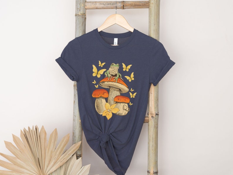 May include: A navy blue t-shirt with a graphic of a green frog sitting on a mushroom with yellow flowers and butterflies.