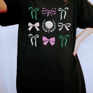 May include: Black t-shirt featuring a golf-themed design. The design includes a golf ball on a tee, surrounded by a wreath, and various bows in shades of green, pink, and white. The shirt is a comfortable, casual style.