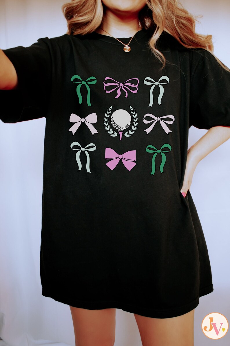 May include: Black t-shirt featuring a golf-themed design. The design includes a golf ball on a tee, surrounded by a wreath, and various bows in shades of green, pink, and white. The shirt is a comfortable, casual style.