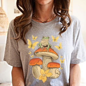 May include: Gray t-shirt with a graphic of a green frog sitting on a mushroom with yellow butterflies.