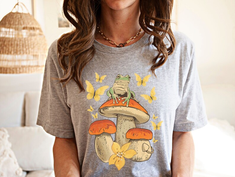 May include: Gray t-shirt with a graphic of a green frog sitting on a mushroom with yellow butterflies.