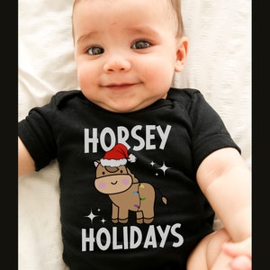 Peut inclure: Un b&eacute;b&eacute; portant un body noir avec un motif blanc repr&eacute;sentant un cheval brun portant un bonnet de P&egrave;re No&euml;l et le texte "HORSEY HOLIDAYS".