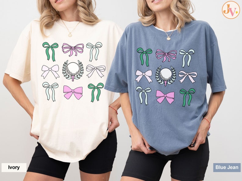 May include: Two oversized t-shirts, one ivory and one blue jean, each featuring a golf-themed design. The design includes a golf ball, a wreath, and various bows in green, pink, and white. The shirts are casual and comfortable.