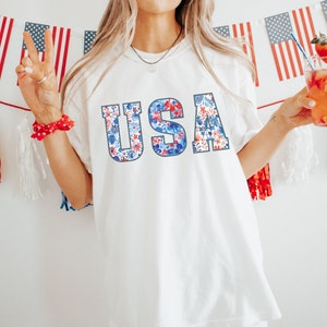 May include: A white t-shirt with a floral print design spelling out "USA" in large letters. The shirt is worn by a person holding a glass of red liquid with a strawberry garnish.