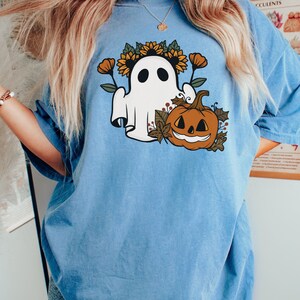 K&ouml;nnte beinhalten: Hellblaues T-Shirt mit einem Halloween-Motiv. Das Design zeigt einen wei&szlig;en Geist mit Sonnenblumenkranz, einen geschnitzten K&uuml;rbis und Herbstlaub. Das T-Shirt hat kurze &Auml;rmel und eine lockere Passform.