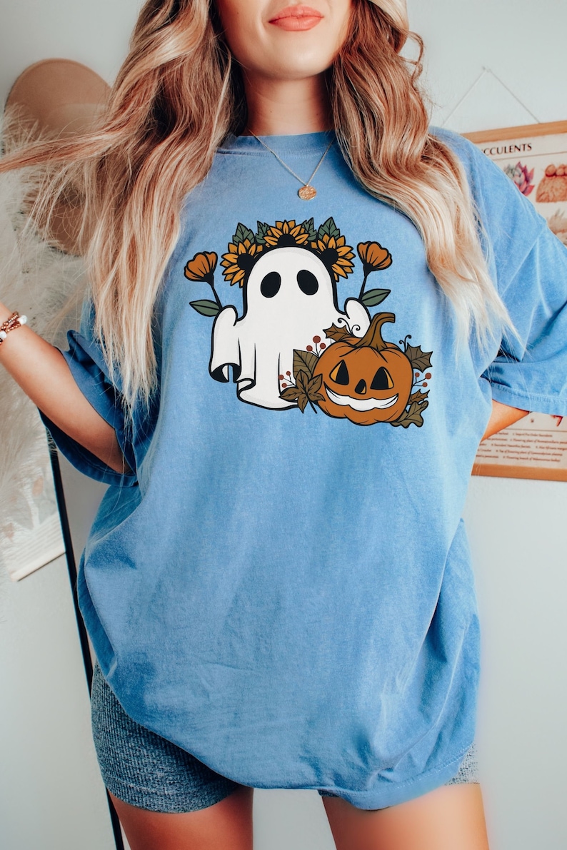 K&ouml;nnte beinhalten: Hellblaues T-Shirt mit einem Halloween-Motiv. Das Design zeigt einen wei&szlig;en Geist mit Sonnenblumenkranz, einen geschnitzten K&uuml;rbis und Herbstlaub. Das T-Shirt hat kurze &Auml;rmel und eine lockere Passform.