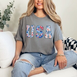 May include: A gray t-shirt with the word "USA" printed on it in large letters. The letters are filled with a red, white, and blue floral pattern.