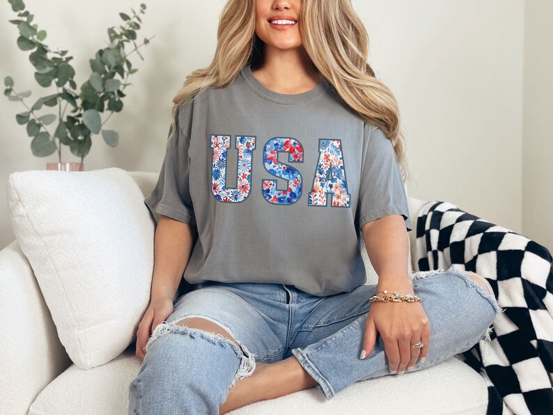 May include: A gray t-shirt with the word "USA" printed on it in large letters. The letters are filled with a red, white, and blue floral pattern.