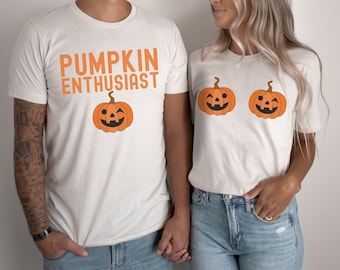May include: A couple wearing white t-shirts with pumpkin designs. The man's shirt says "Pumpkin Enthusiast" and has a single pumpkin with a winking face. The woman's shirt has two pumpkins with winking faces.