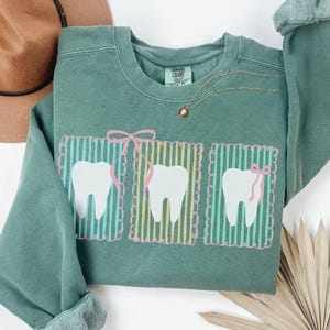 May include: A light green sweatshirt with a graphic of three white teeth, each framed by vertical stripes and a pink bow. The sweatshirt has a crew neck and long sleeves. A gold necklace with a small pendant is visible.