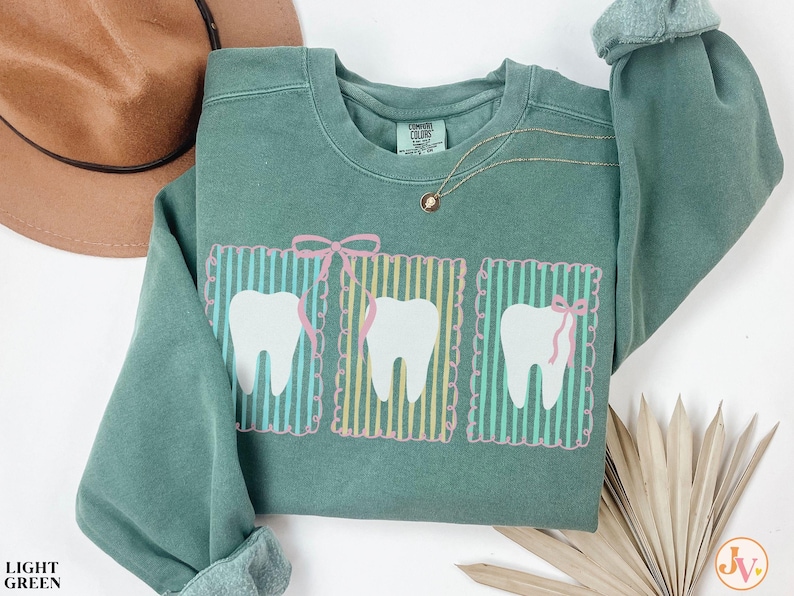 May include: A light green sweatshirt with a graphic of three white teeth, each framed by vertical stripes and a pink bow. The sweatshirt has a crew neck and long sleeves. A gold necklace with a small pendant is visible.