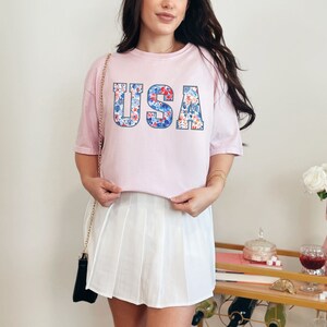 May include: A pink t-shirt with a floral print "USA" graphic. The shirt is worn with a white pleated skirt.