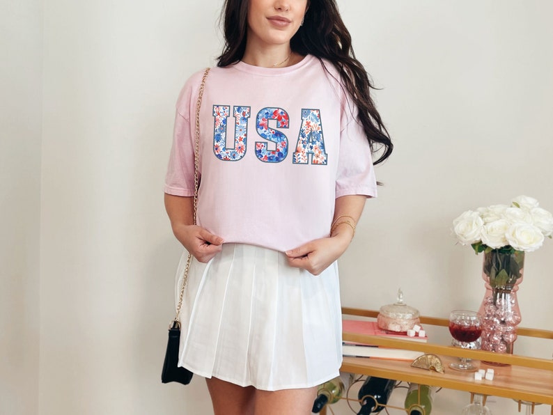 May include: A pink t-shirt with a floral print "USA" graphic. The shirt is worn with a white pleated skirt.