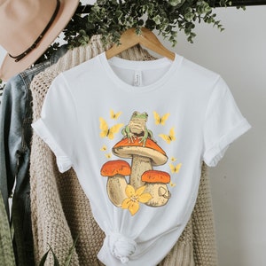 May include: White t-shirt with a graphic of a green frog sitting on a mushroom with yellow butterflies. The mushroom is orange and brown with a yellow flower.