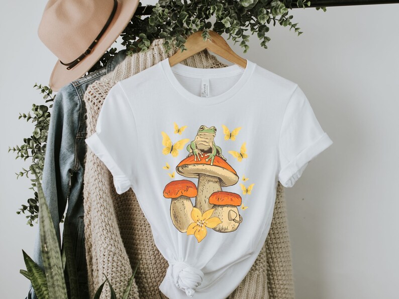 May include: White t-shirt with a graphic of a green frog sitting on a mushroom with yellow butterflies. The mushroom is orange and brown with a yellow flower.
