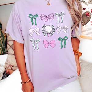 May include: A light purple t-shirt featuring a golf-themed design. The design includes a golf ball on a tee surrounded by a laurel wreath, along with various bows in shades of pink, green, and white.