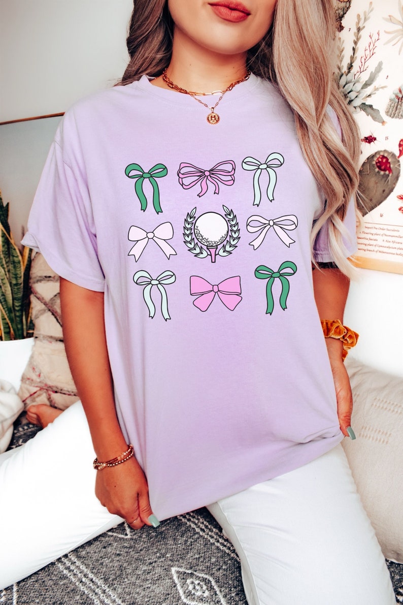 May include: A light purple t-shirt featuring a golf-themed design. The design includes a golf ball on a tee surrounded by a laurel wreath, along with various bows in shades of pink, green, and white.