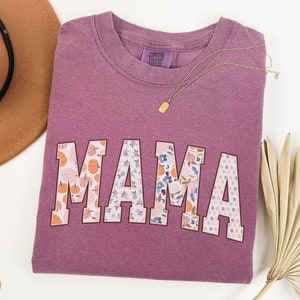 May include: A purple t-shirt with the word "MAMA" in large, white letters. Each letter is decorated with a different floral and pumpkin pattern.
