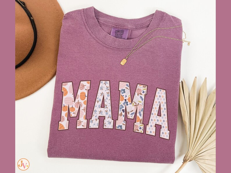 May include: A purple t-shirt with the word "MAMA" in large, white letters. Each letter is decorated with a different floral and pumpkin pattern.
