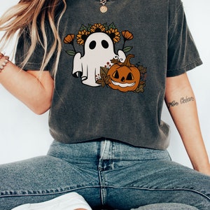 K&ouml;nnte beinhalten: Dunkelgraues T-Shirt mit einem Halloween-Motiv. Das Design zeigt einen wei&szlig;en Geist mit Sonnenblumenkranz und einen l&auml;chelnden K&uuml;rbis. Das T-Shirt wird mit blauen Jeans und wei&szlig;en Turnschuhen getragen.