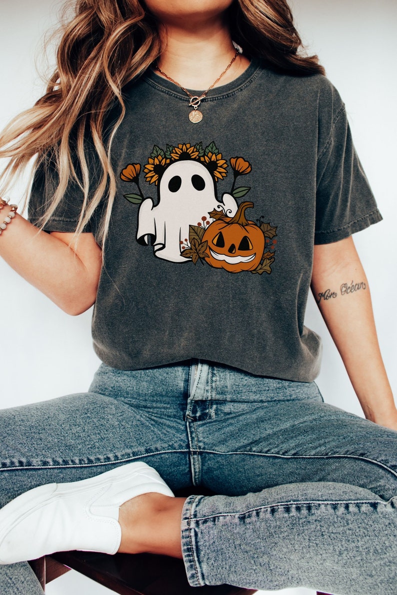 K&ouml;nnte beinhalten: Dunkelgraues T-Shirt mit einem Halloween-Motiv. Das Design zeigt einen wei&szlig;en Geist mit Sonnenblumenkranz und einen l&auml;chelnden K&uuml;rbis. Das T-Shirt wird mit blauen Jeans und wei&szlig;en Turnschuhen getragen.