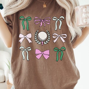 May include: Brown t-shirt featuring a golf-themed design. The design includes a golf ball surrounded by a wreath, along with several colorful bows in shades of green, pink, and light blue. The shirt is a comfortable, casual style.