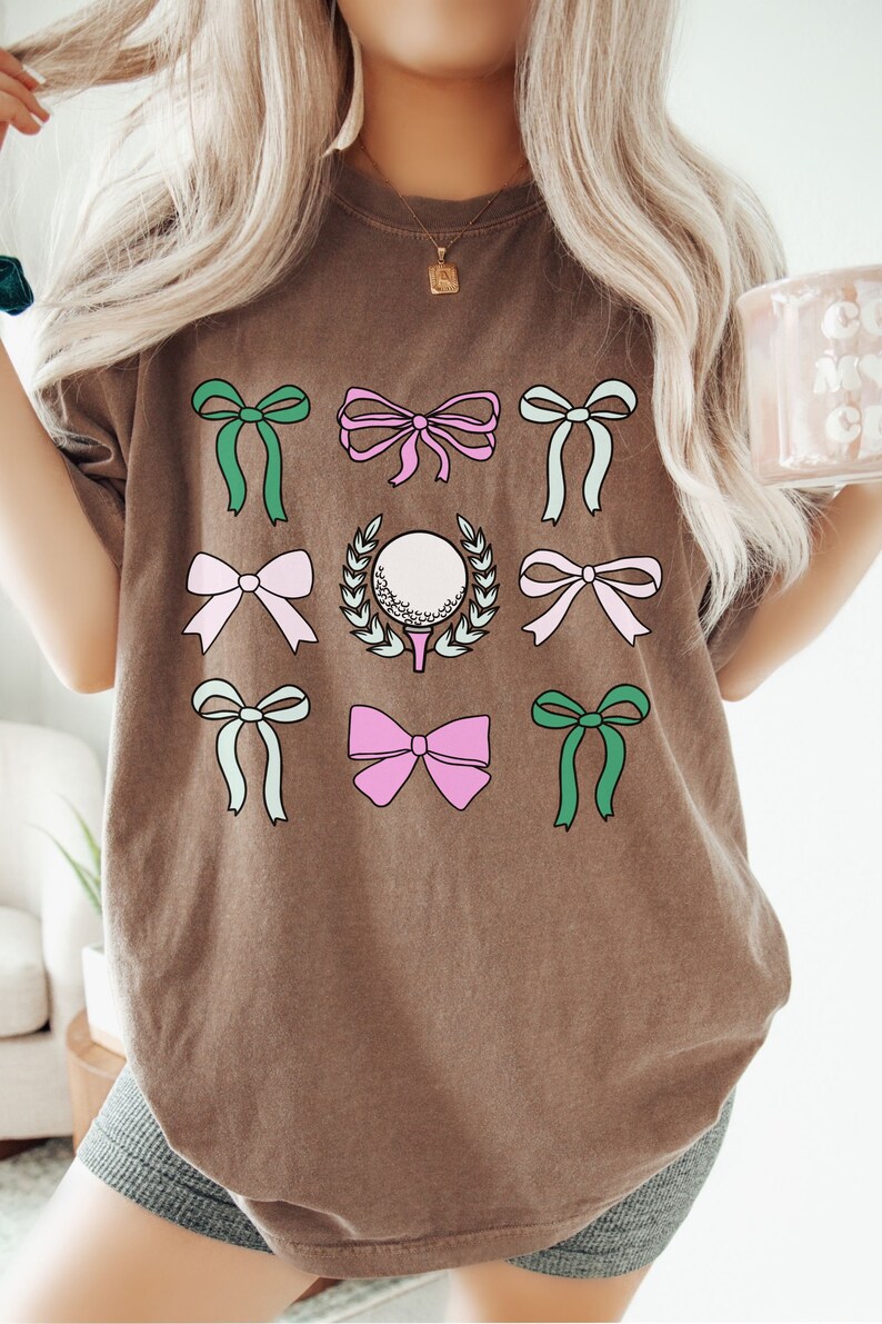 May include: Brown t-shirt featuring a golf-themed design. The design includes a golf ball surrounded by a wreath, along with several colorful bows in shades of green, pink, and light blue. The shirt is a comfortable, casual style.