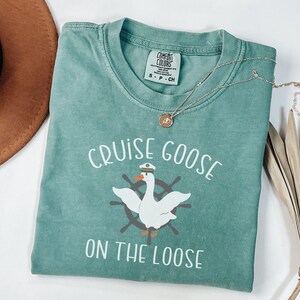 Puede incluir: Una camiseta azul verdosa con el texto "Cruise Goose on the Loose" y un dibujo animado de un ganso con un sombrero de capit&aacute;n y un tim&oacute;n de barco.