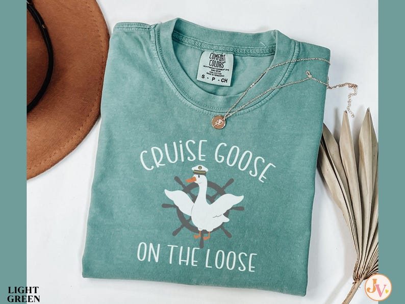 Puede incluir: Una camiseta azul verdosa con el texto "Cruise Goose on the Loose" y un dibujo animado de un ganso con un sombrero de capit&aacute;n y un tim&oacute;n de barco.