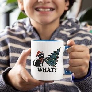 May include: White ceramic mug with a blue rim. The mug features a black cat wearing a Santa hat and scarf, knocking over a decorated Christmas tree. The text "WHAT?" is printed below the image.
