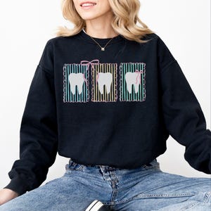 May include: Black sweatshirt featuring three tooth graphics. Each tooth is framed with a colorful striped border and a pink bow or dental floss. The sweatshirt is a comfortable, casual style, suitable for dental professionals or those who appreciate dental-themed designs.