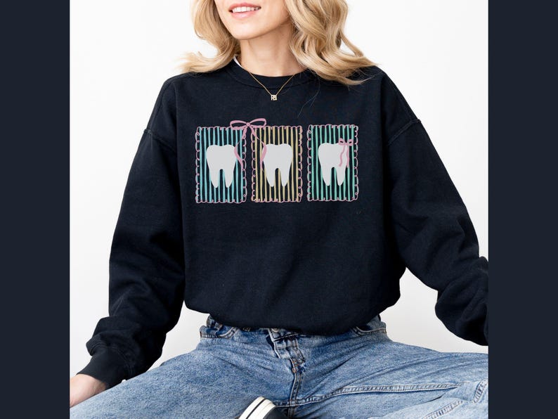 May include: Black sweatshirt featuring three tooth graphics. Each tooth is framed with a colorful striped border and a pink bow or dental floss. The sweatshirt is a comfortable, casual style, suitable for dental professionals or those who appreciate dental-themed designs.