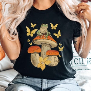 May include: A black t-shirt with a graphic of a green frog sitting on a mushroom with yellow butterflies. The mushroom has a brown stem and a white cap with orange accents.