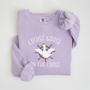 Puede incluir: Una sudadera de color lavanda con el texto "Cruise Goose on the Loose" y un dibujo animado de un ganso con un sombrero de capit&aacute;n y un tim&oacute;n de barco.