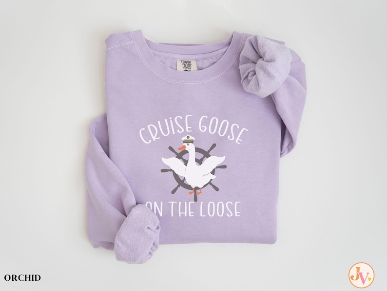 Puede incluir: Una sudadera de color lavanda con el texto "Cruise Goose on the Loose" y un dibujo animado de un ganso con un sombrero de capit&aacute;n y un tim&oacute;n de barco.